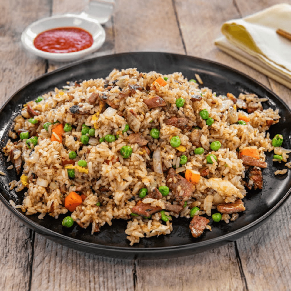 Steaming fried rice with peas, carrots, and meat on a black plate, with a side of red sauce.