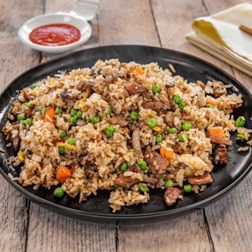 Steaming fried rice with peas, carrots, and meat on a black plate, with a side of red sauce.