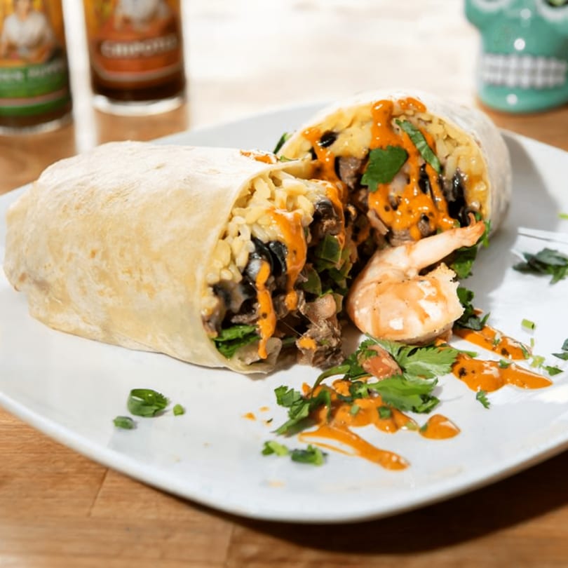 A Nara Teriyaki and Pho burrito, cut in half, with shrimp, sauce, and cilantro on a white plate.